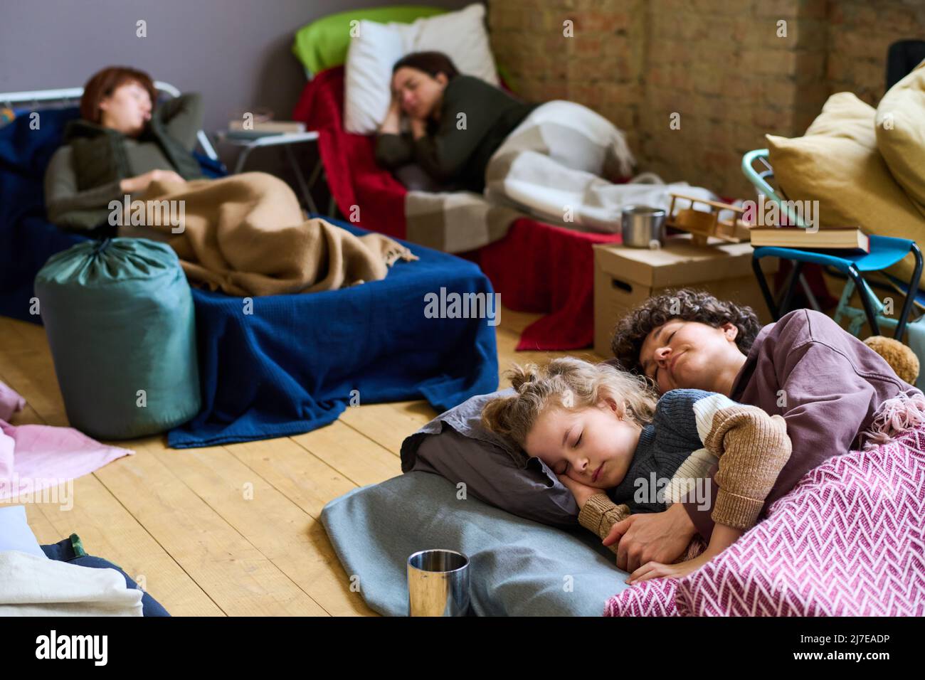 Giovane donna pacifica e suo figlio carino che dormono sul materasso sul pavimento o ampio spazio contro altre donne rifugiati Foto Stock