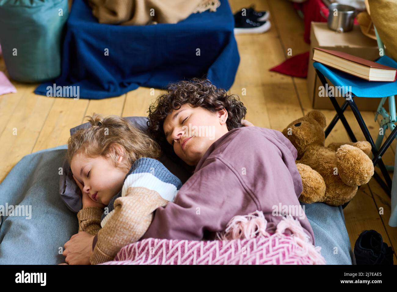 Adorabile ragazzo con capelli biondi ricci e sua madre che dorme sotto la coperta sul pavimento della stanza con i materassi per i rifugiati Foto Stock