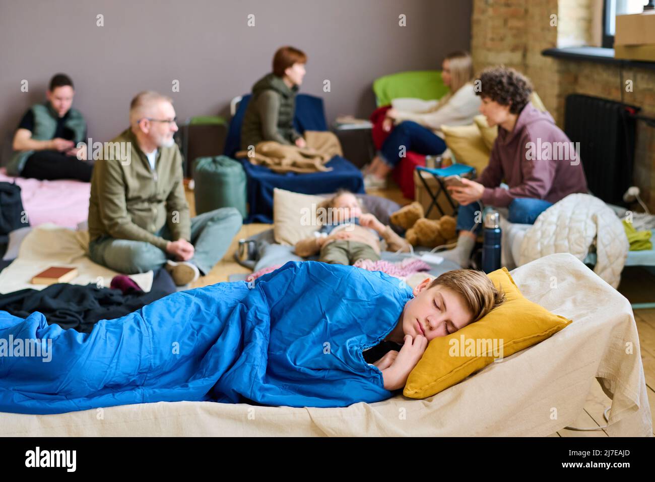 Ragazzo adolescente che tiene la testa su cuscino mentre dorme sotto coperta blu contro gruppo di persone che comunicano sui loro letti Foto Stock