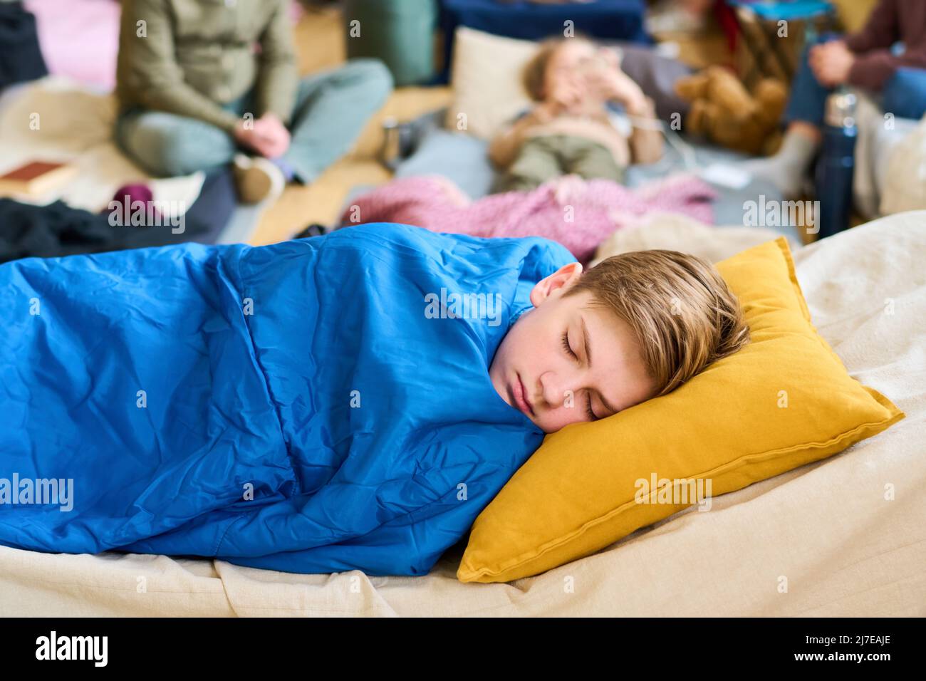 Un ragazzo tranquillo che napping sul posto letto sotto la coperta blu mentre tiene la testa su cuscino giallo contro il gruppo di rifugiati Foto Stock