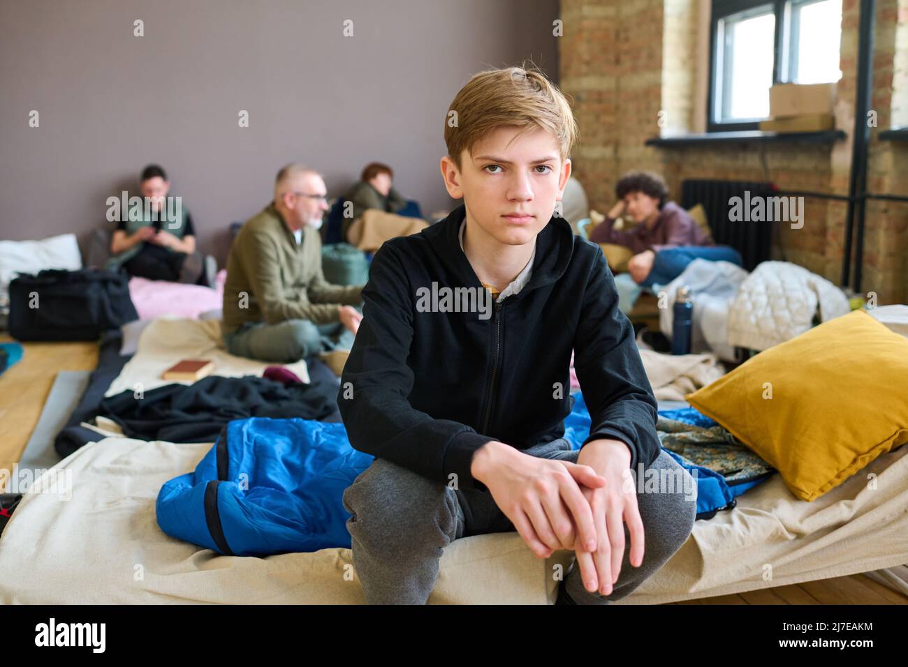 Ragazzo serio in casualwear seduto su cuchette preparato per i senzatetto e guardando la macchina fotografica contro i rifugiati che parlano tra loro Foto Stock