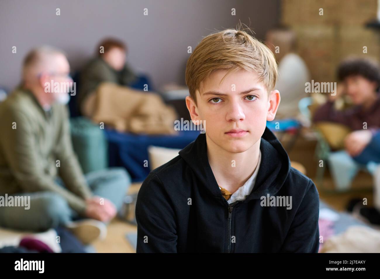 Un ragazzo serio che guarda la macchina fotografica mentre è seduto contro altri rifugiati e temporaneamente senzatetto che comunicano tra loro Foto Stock