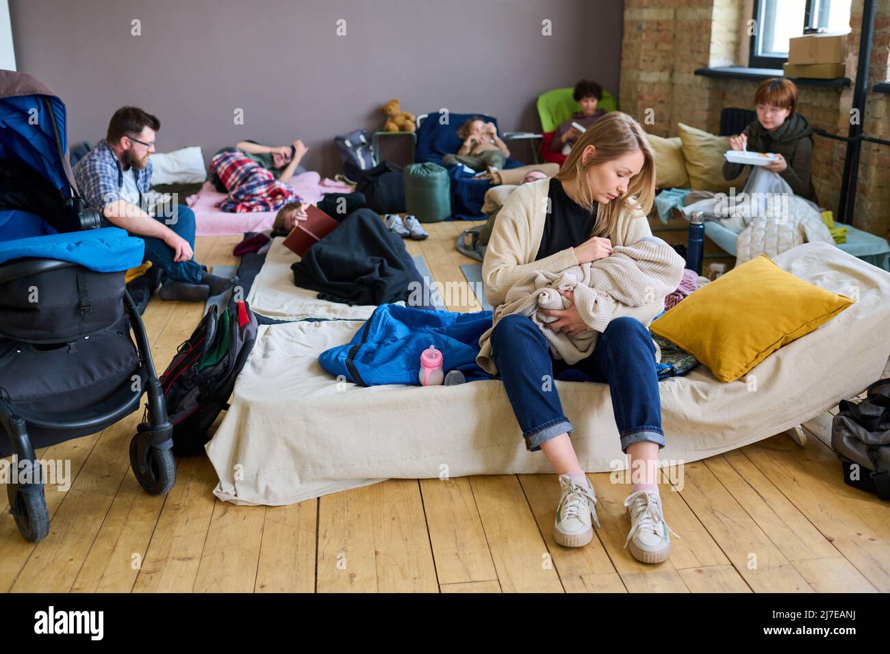 Giovane mamma attenta in casualwear lulling il suo bambino avvolto in un caldo plaid mentre si siede sul posto di sonno in rifugio per i rifugiati Foto Stock