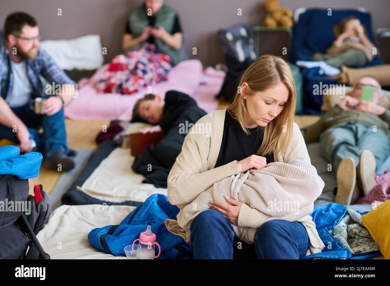 Giovane donna bionda rifugiata lulling bambino avvolto in caldo plaid mentre si siede a letto preparato da volontari per le persone in difficoltà Foto Stock