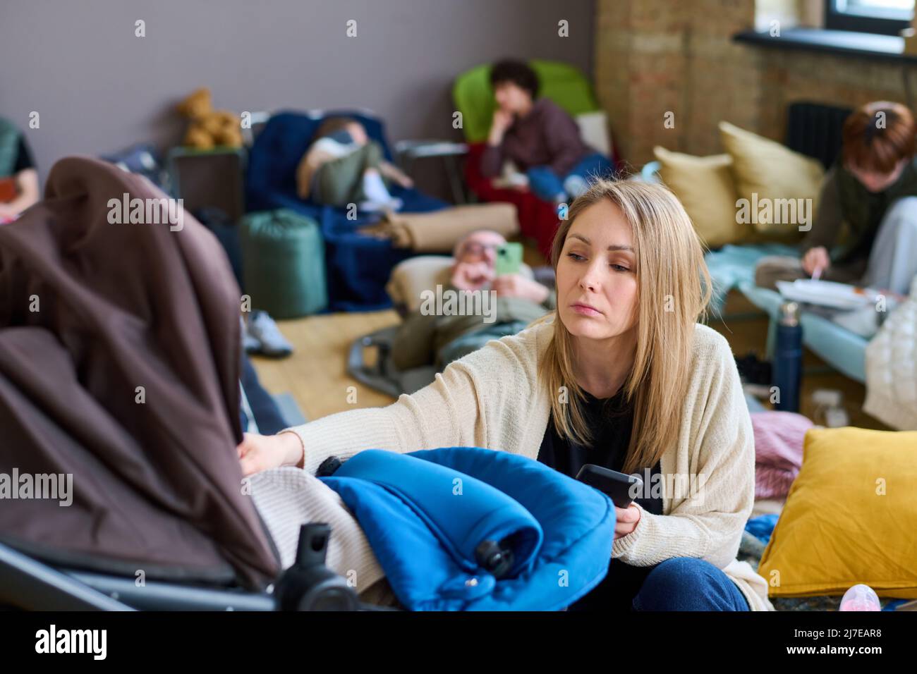 Giovane mamma attenta lulling suo bambino in passeggino mentre si siede sul posto letto in camera spaziosa con temporaneamente senzatetto persone Foto Stock