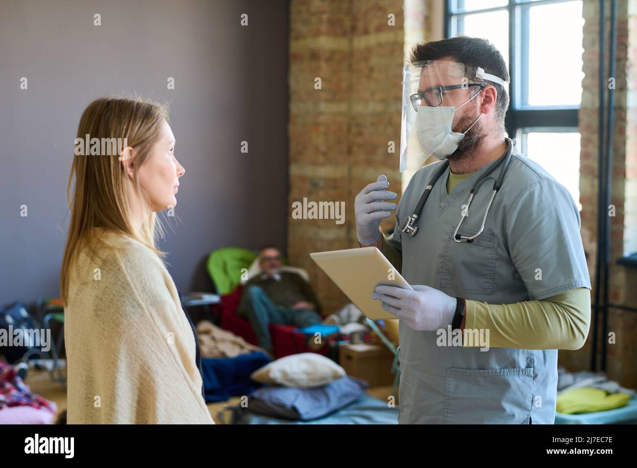 Medico di sesso maschile con tavoletta che dà consigli medici alla giovane rifugiata di sesso femminile malata nel campo e spiega come trattare la sua malattia Foto Stock