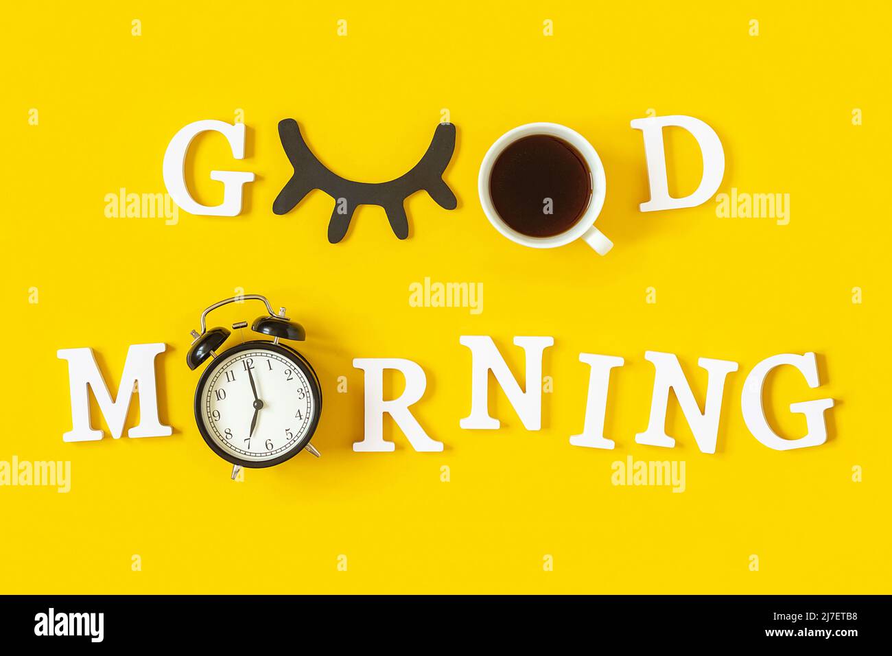 Concetto creativo di buon mattino. Lettere bianche, ciglia nere, sveglia e tazza di caffè su sfondo giallo. Vista dall'alto, disposizione piatta. Biglietto di auguri. Foto Stock