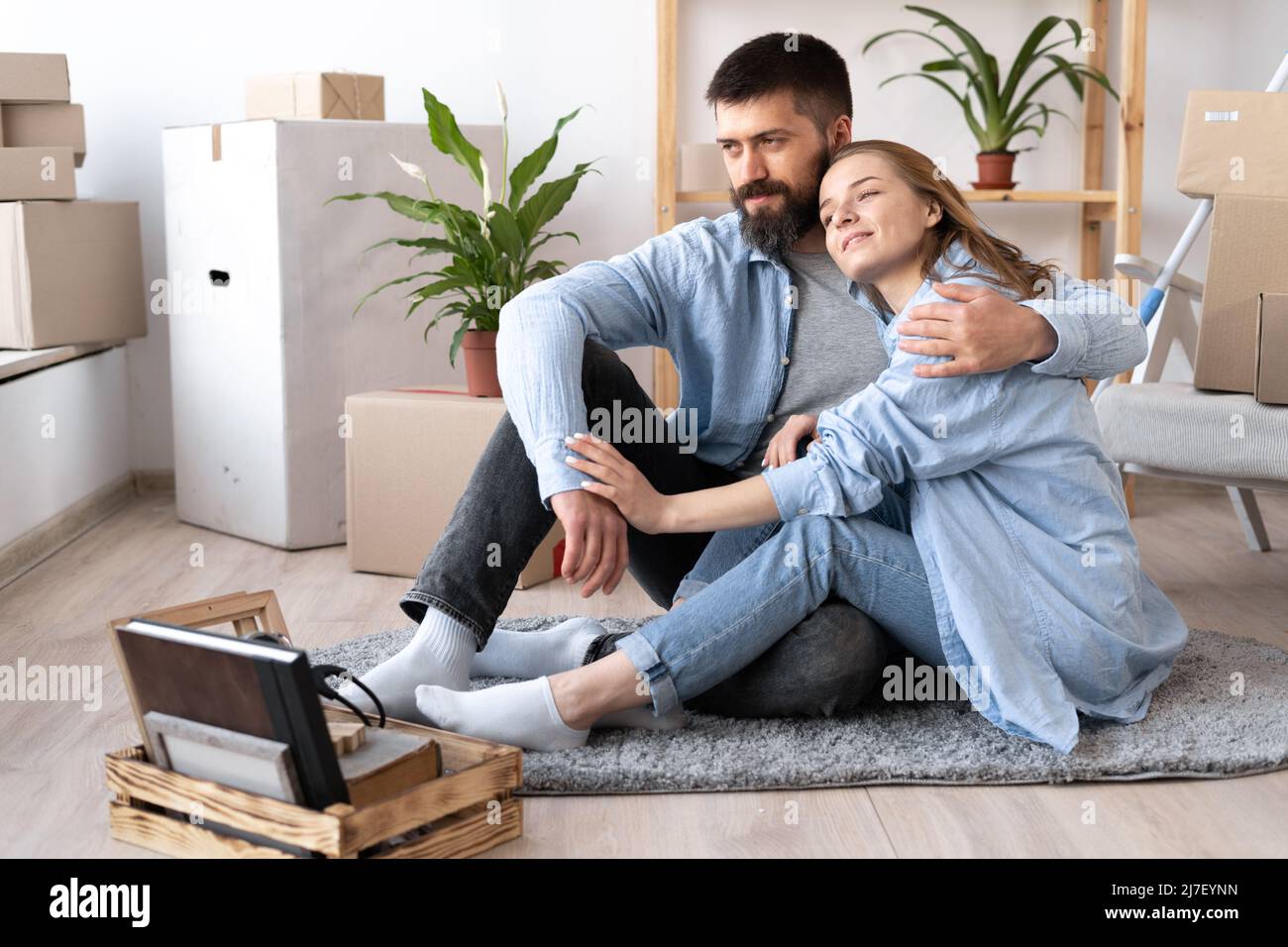 felice giovane coppia uomo e donna sono seduti sul pavimento in abiti casual e abbraccio. sposta scatole di cartone e mobili durante un passaggio a un nuovo Foto Stock