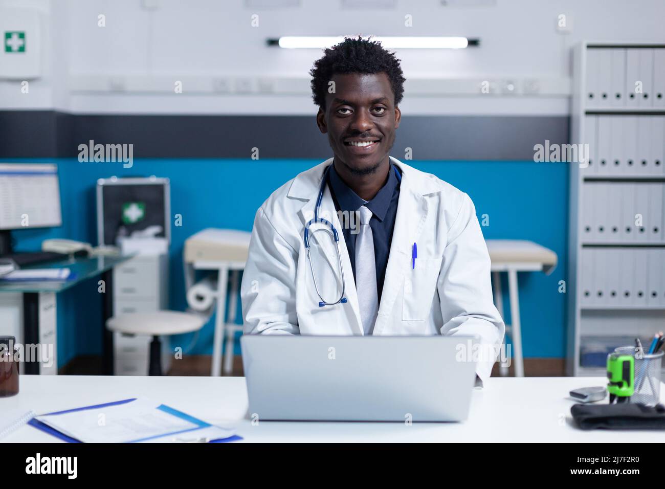 Sorridente specialista di clinica sanitaria afroamericana seduto alla scrivania mentre si utilizza un computer portatile. Medico ospedaliero che indossa un cappotto bianco uniforme seduto alla scrivania nello spazio di lavoro mentre si utilizza il computer moderno Foto Stock