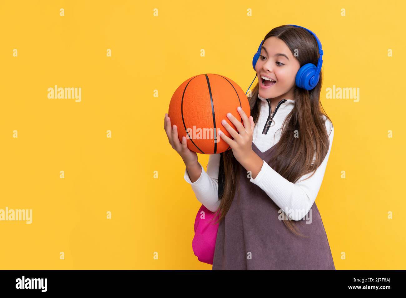 il bambino ha scioccato in cuffie con zaino scuola e palla da basket su sfondo giallo Foto Stock
