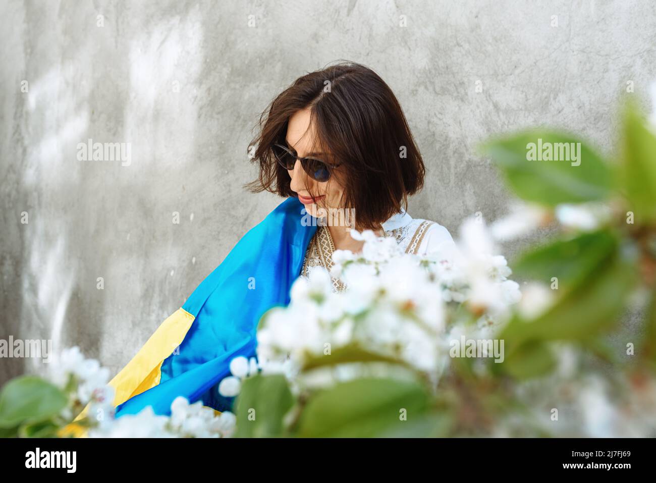 Ragazza in un abito bianco e occhiali da sole su uno sfondo di muro di cemento detiene la bandiera nazionale che celebra la vittoria. Fiori di mela e pera in primo piano. Spazio di copia. Giorno della Costituzione Foto Stock