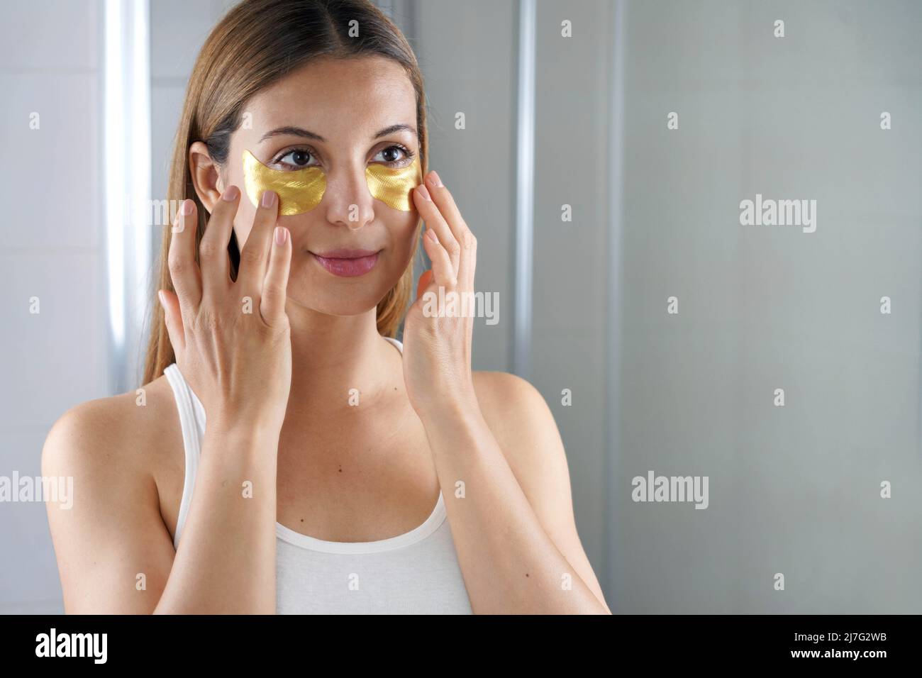 Donna di bellezza che applica maschera sotto-occhio dorata anti-invecchiamento che guarda se stessa nello specchio nel bagno. Cura della pelle ragazza toccare patch di tessuto maschera unde Foto Stock