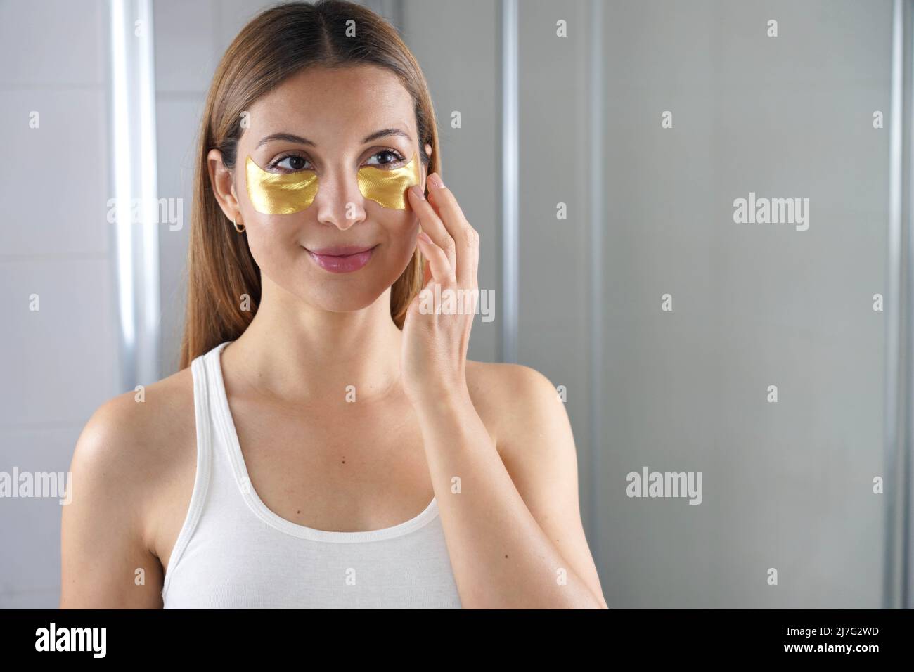 Donna di bellezza che applica maschera sotto-occhio dorata anti-invecchiamento che guarda se stessa nello specchio nel bagno. Cura della pelle ragazza toccare patch di tessuto maschera unde Foto Stock