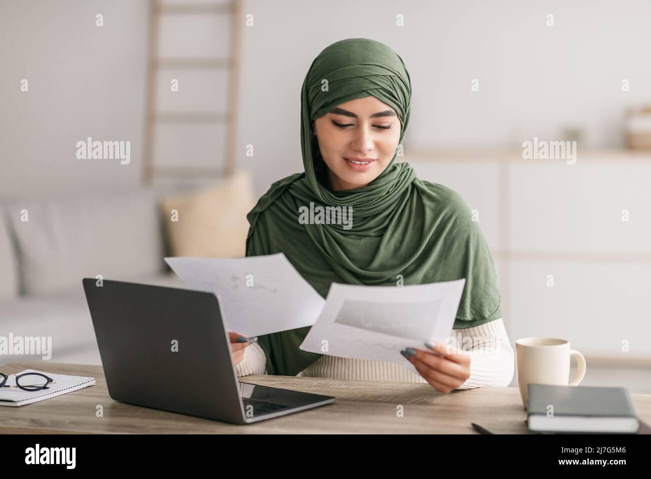 Felice giovane donna araba in hijab usando il laptop, guardando attraverso documenti di lavoro durante la riunione di lavoro in linea a casa Foto Stock