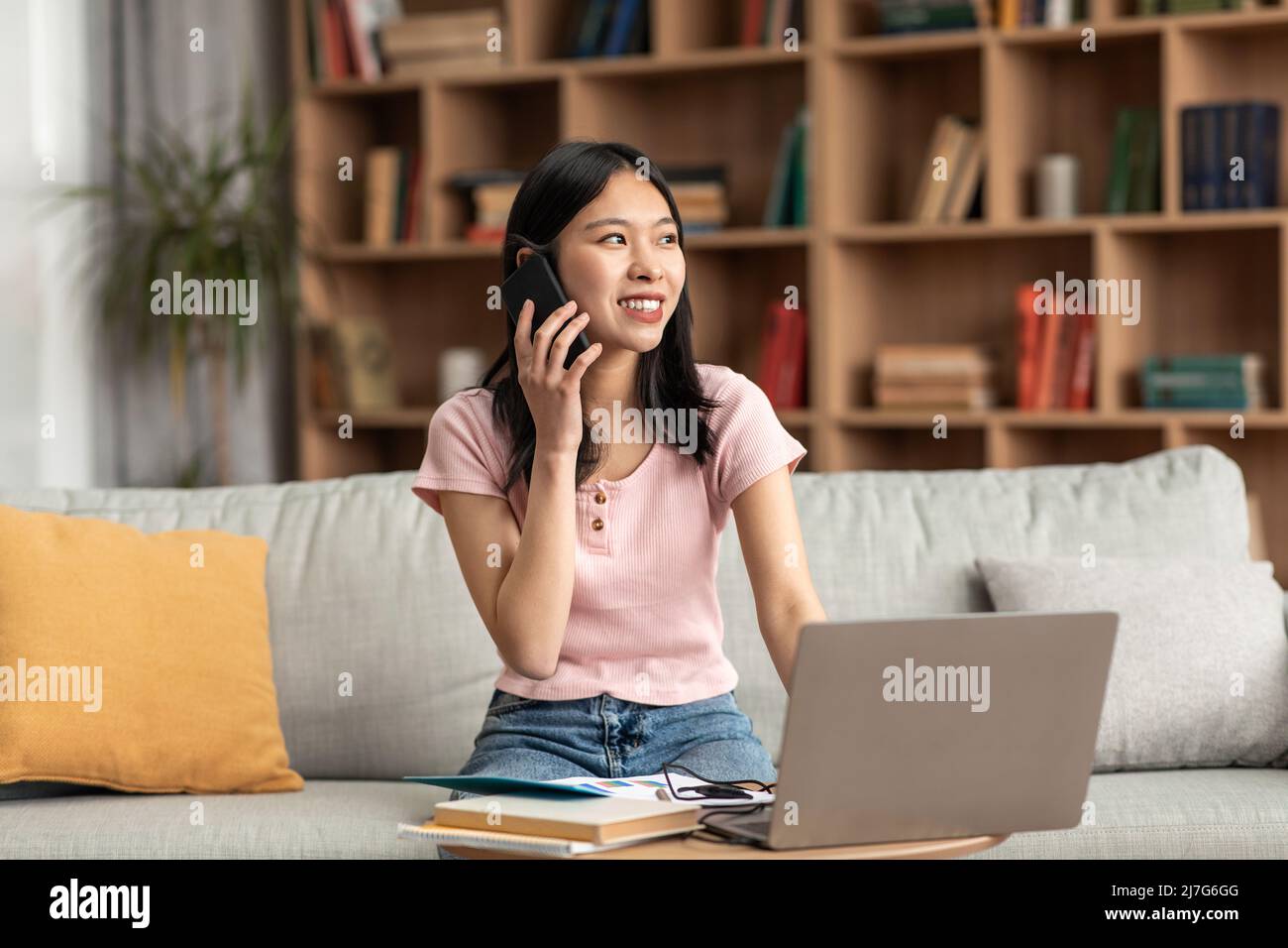 Signora coreana positiva che ha una conversazione telefonica mentre si siede sul divano a casa e usando il portatile, spazio copia Foto Stock