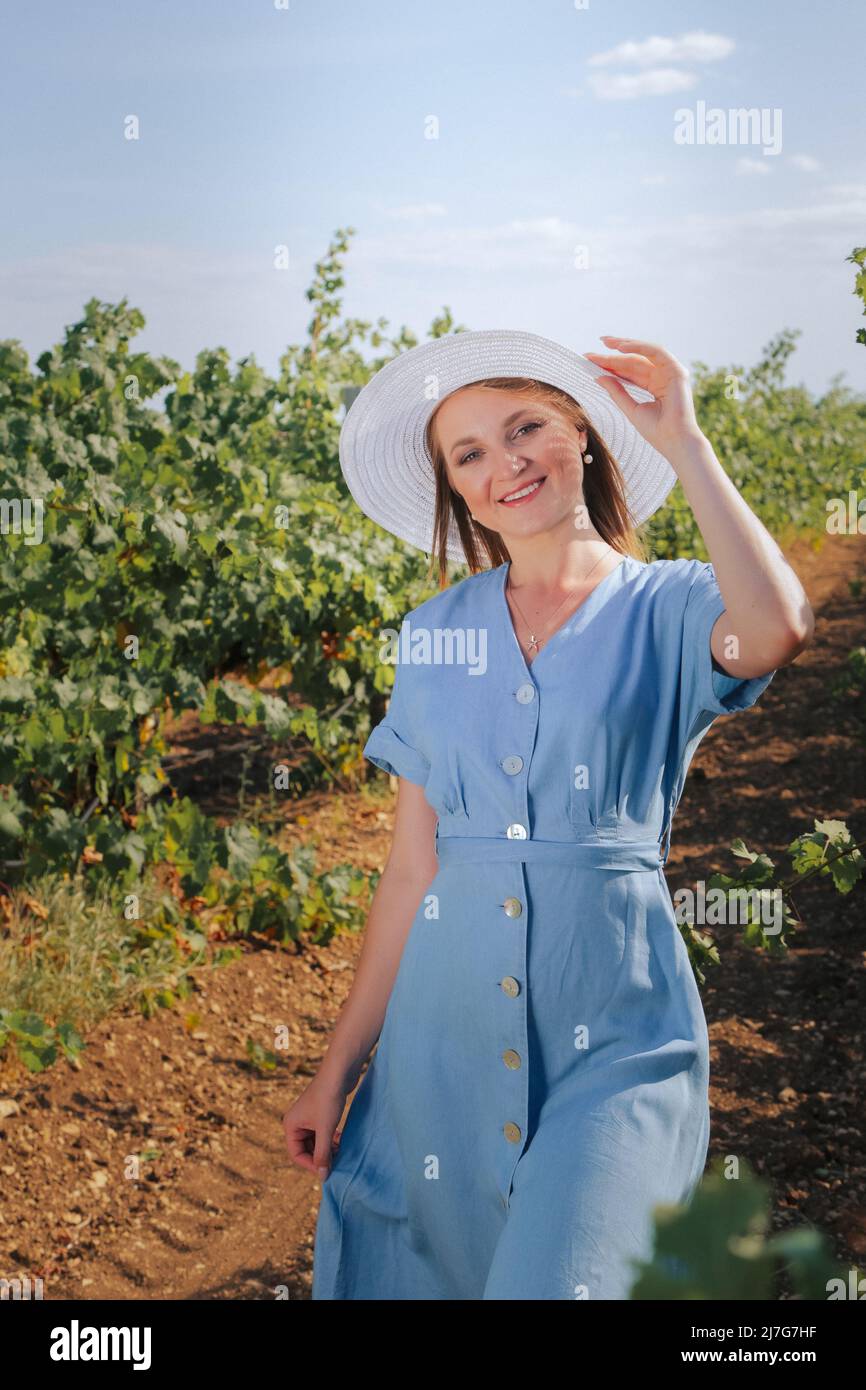 Una ragazza bionda in un vigneto, in piedi tra le file di alberi e sorridendo in un abito blu di nazionalità caucasica Foto Stock