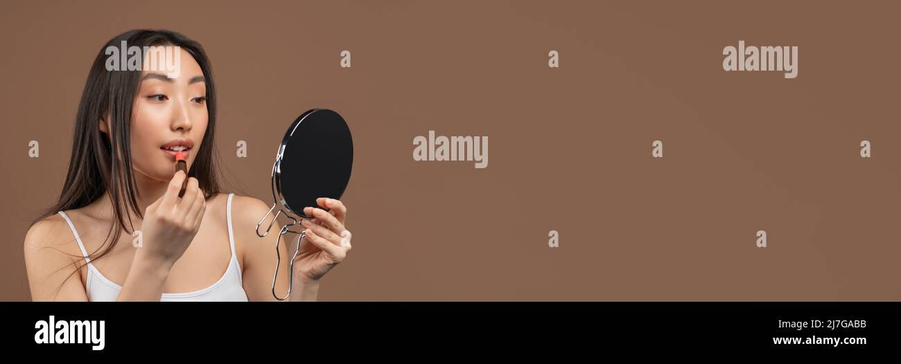 Graziosa signora asiatica che fa il trucco quotidiano, usando specchio e rossetto su sfondo marrone studio, panorama con spazio libero Foto Stock