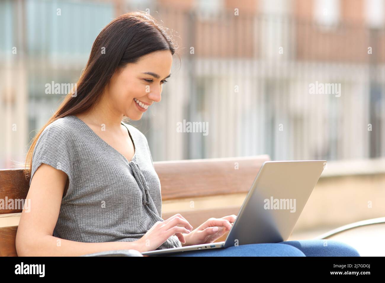 Donna felice sorridendo usando il portatile seduto sulla panca in strada Foto Stock