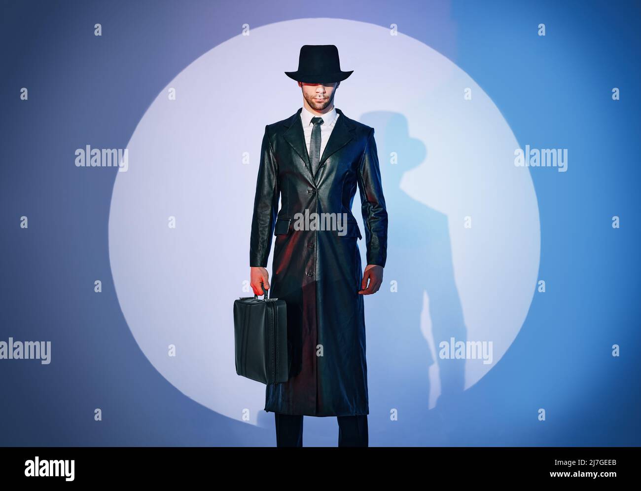 Ritratto a tutta lunghezza di un bell'uomo in cappotto nero e portadocumenti con cappello che si posa in primo piano sullo sfondo dello studio. stile film noir. Privato de Foto Stock