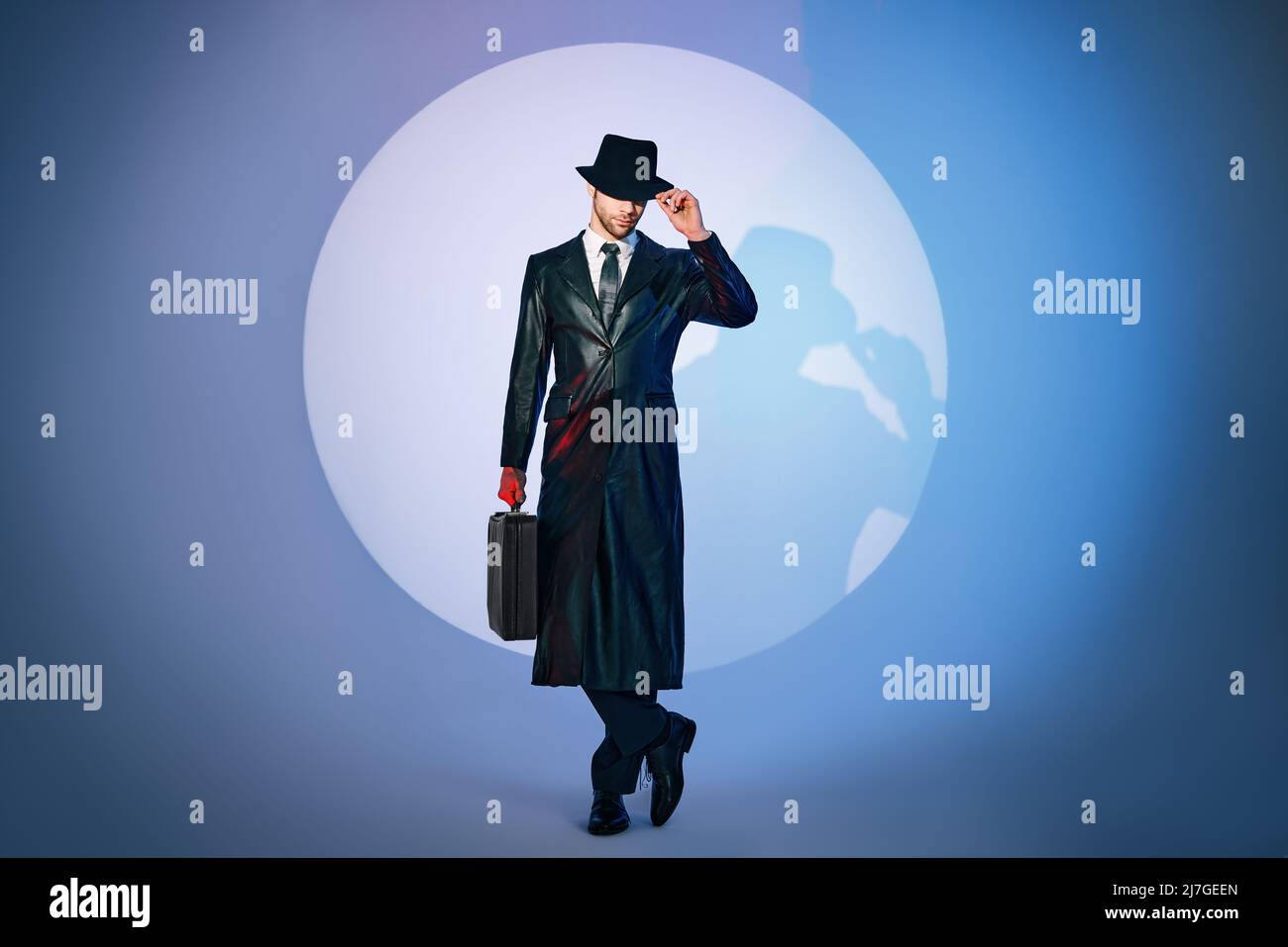 Ritratto a tutta lunghezza di un bell'uomo in cappotto nero e portadocumenti con cappello che si posa in primo piano sullo sfondo dello studio. stile film noir. Privato de Foto Stock