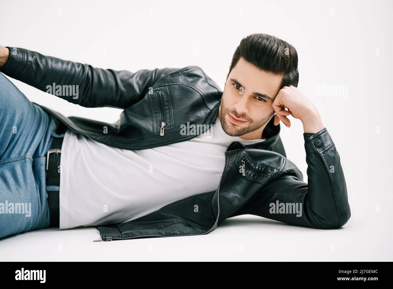 Bell'uomo elegante che si posa sul pavimento su sfondo bianco studio. Concetto di bellezza maschile. Giovane uomo in jeans, t-shirt bianca e pelle nera ja Foto Stock