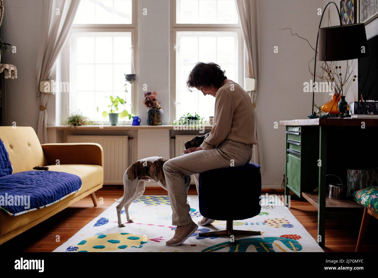 Vista laterale della donna anziana che accarezzava il cane mentre si siede sullo sgabello ottomano nel soggiorno di casa Foto Stock