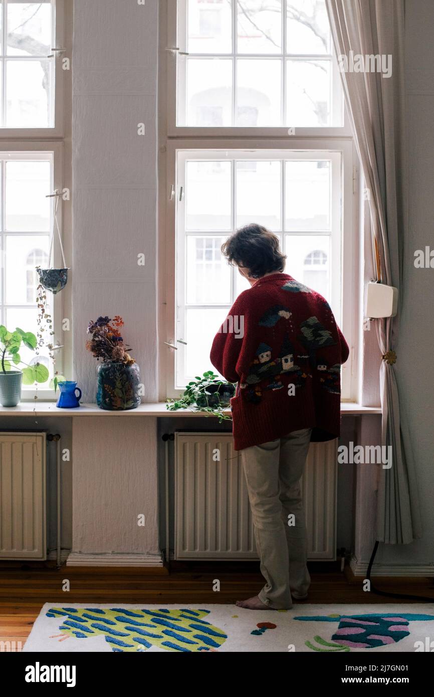 Vista posteriore della donna anziana in piedi vicino alla finestra nel soggiorno di casa Foto Stock