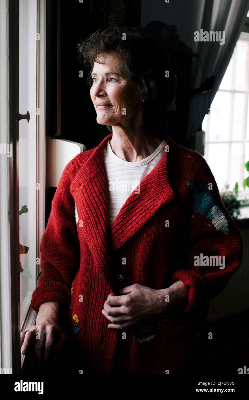 Donna anziana pensierosa sorridente mentre guarda attraverso la finestra a casa Foto Stock