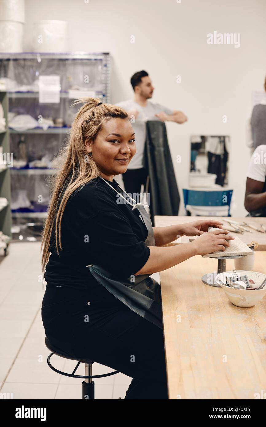Ritratto di giovane donna sorridente con capelli biondi lunghi che modellano argilla mentre si siede a tavola in classe d'arte Foto Stock