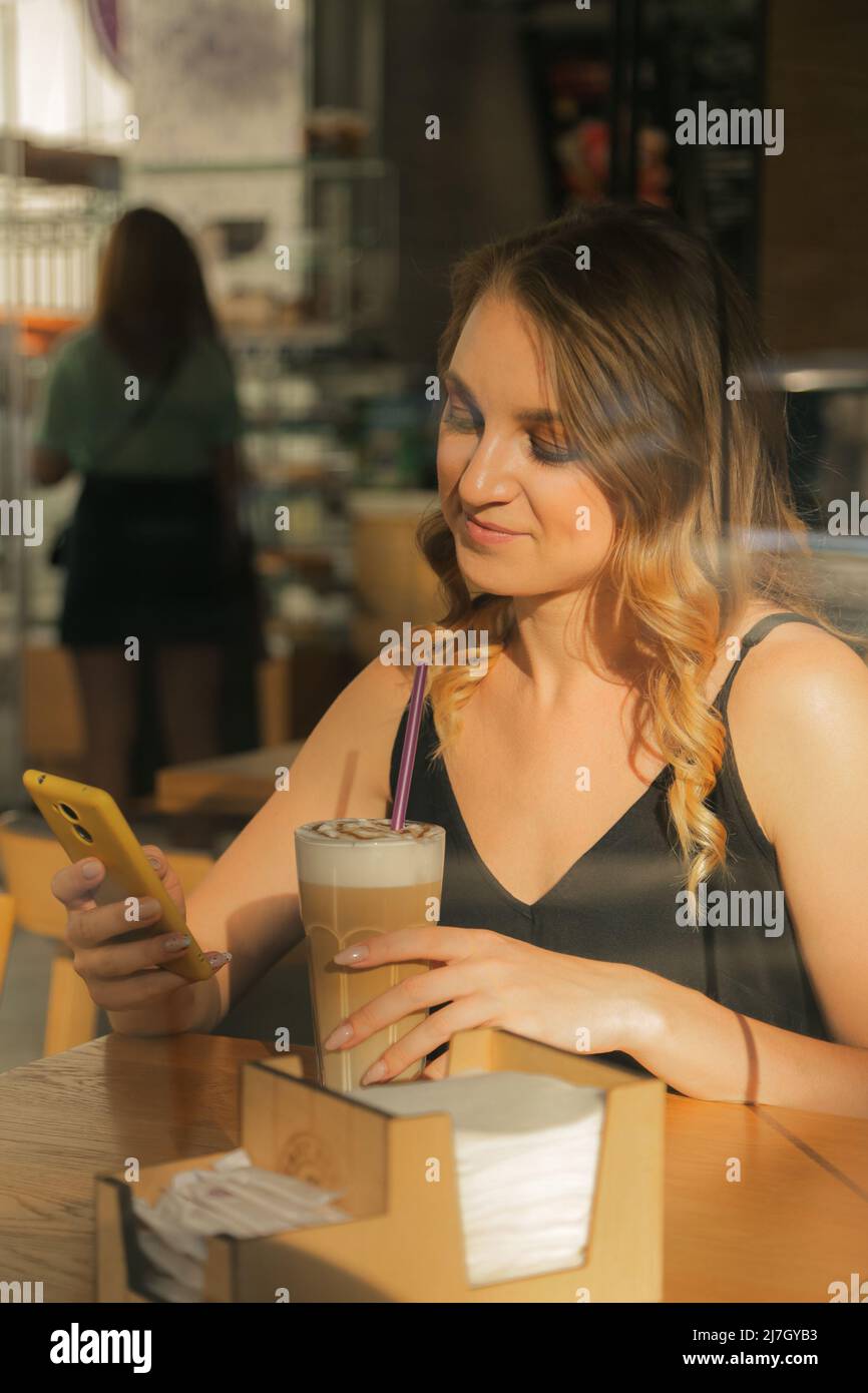 Donna d'affari seduta in un caffè, SMS su uno smartphone, vista attraverso la finestra Foto Stock