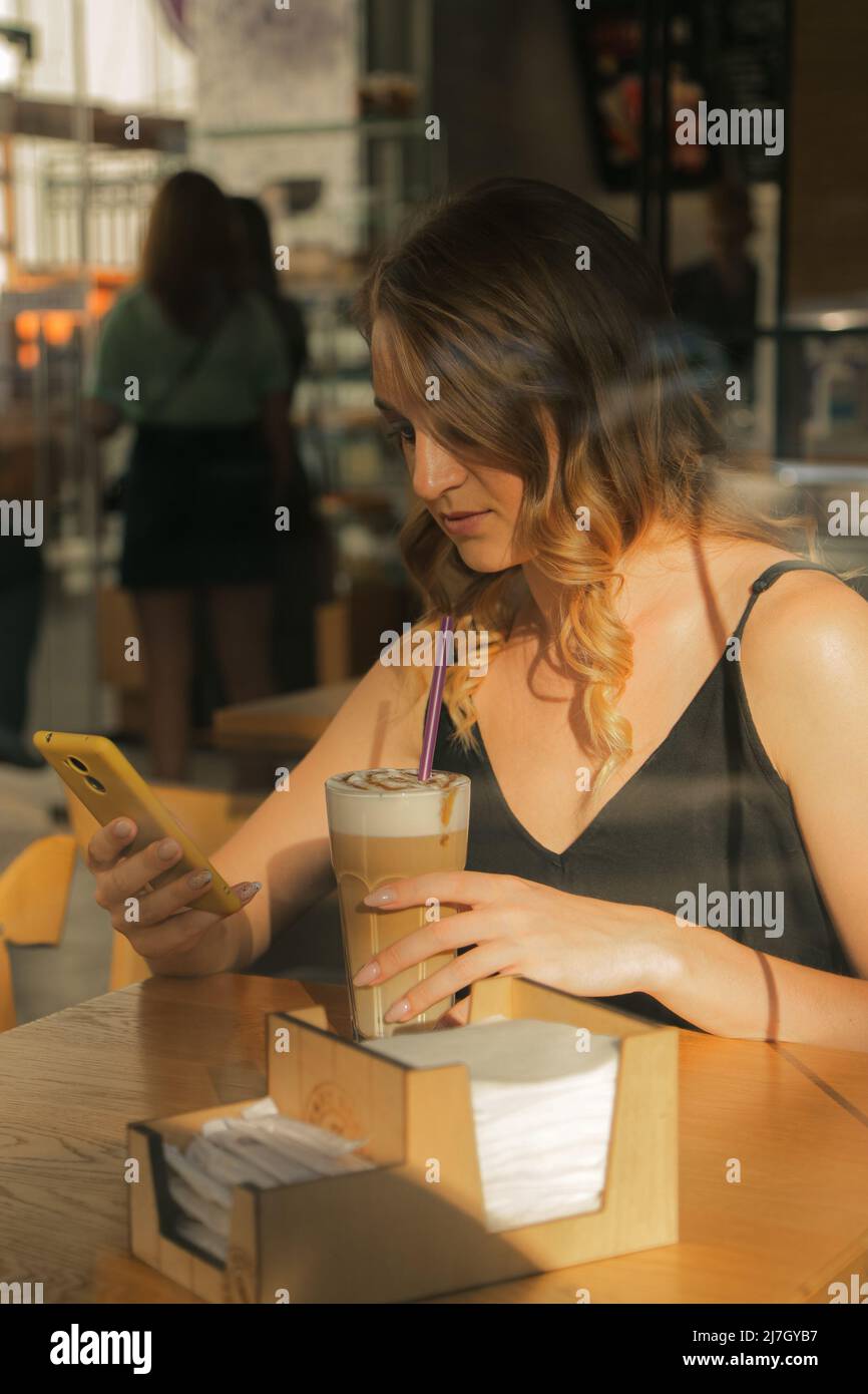 Una donna piuttosto giovane beve caffè in un caffè e chiacchiera su uno smartphone, vista attraverso il vetro Foto Stock