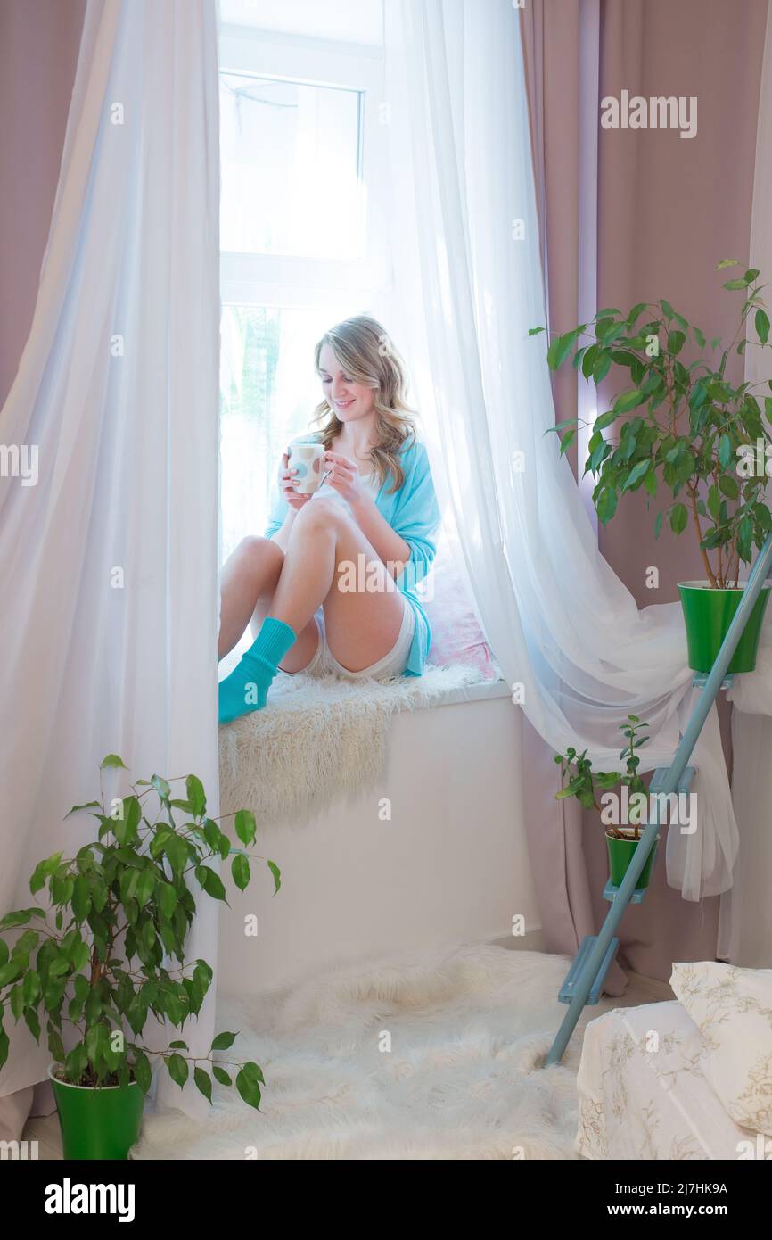 Una ragazza gioiosa inizia la sua mattina con una tazza di tè o caffè su un accogliente davanzale nella sua luminosa camera da letto Foto Stock
