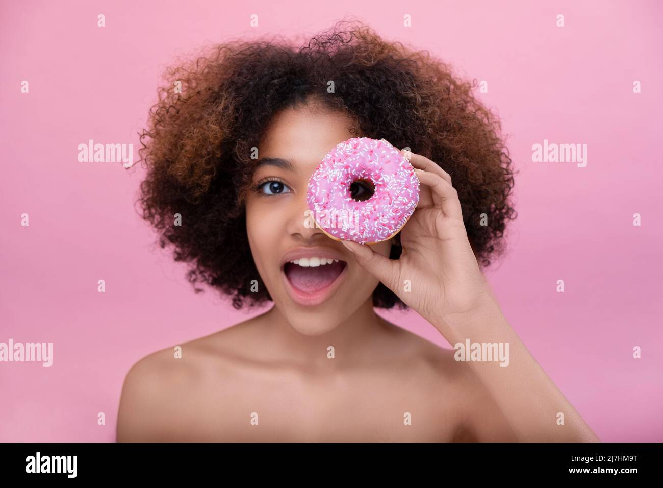 Ritratto di una giovane donna felice con pelle scura copre un occhio con una ciambella rosa su sfondo rosa, spazio copia Foto Stock