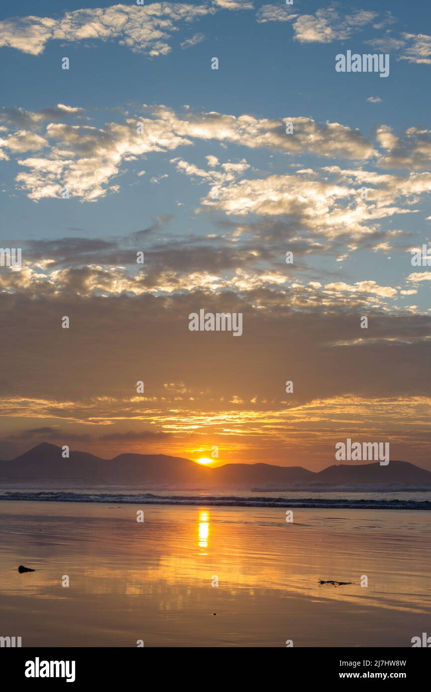 Al tramonto sulla spiaggia di Famara, i raggi del sole illuminano le nuvole in rosso e oro, che si riflettono nella sabbia bagnata, silhouette delle montagne. Foto Stock