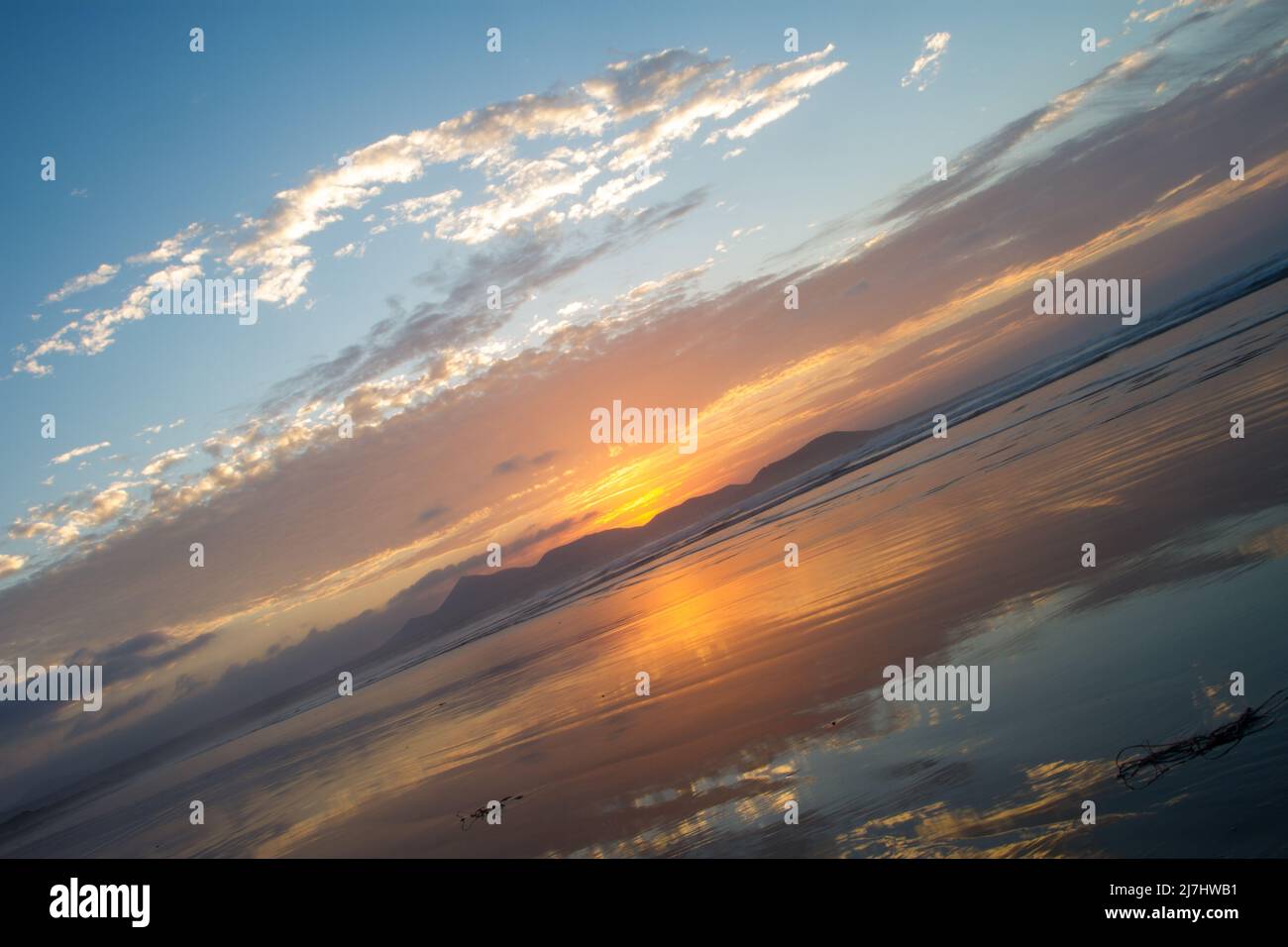 Al tramonto sulla spiaggia di Famara, i raggi del sole illuminano le nuvole in rosso e oro, che si riflettono nella sabbia bagnata, silhouette delle montagne. Foto Stock