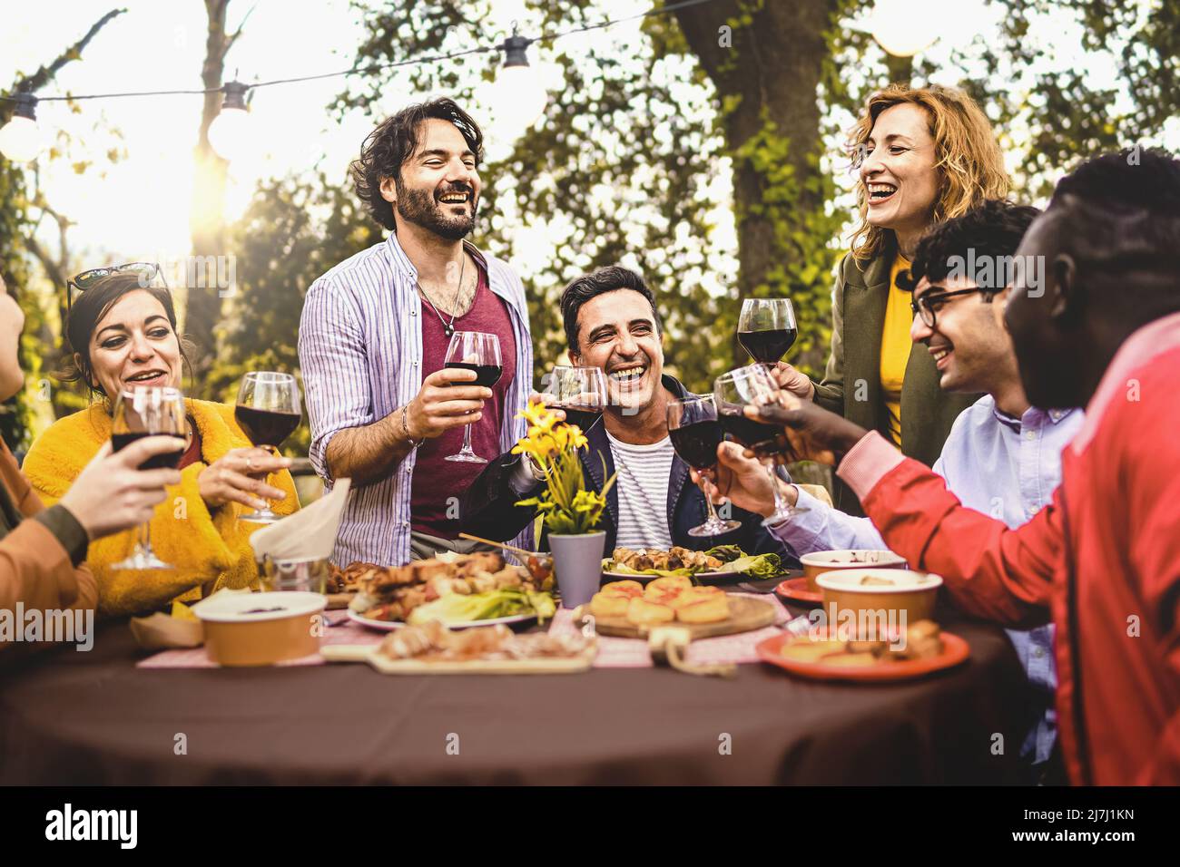 Gruppo di amici che cenano all'aperto tostando con vino rosso nel cortile - persone diverse di varie generazioni che si divertono insieme nella natura seduta Foto Stock