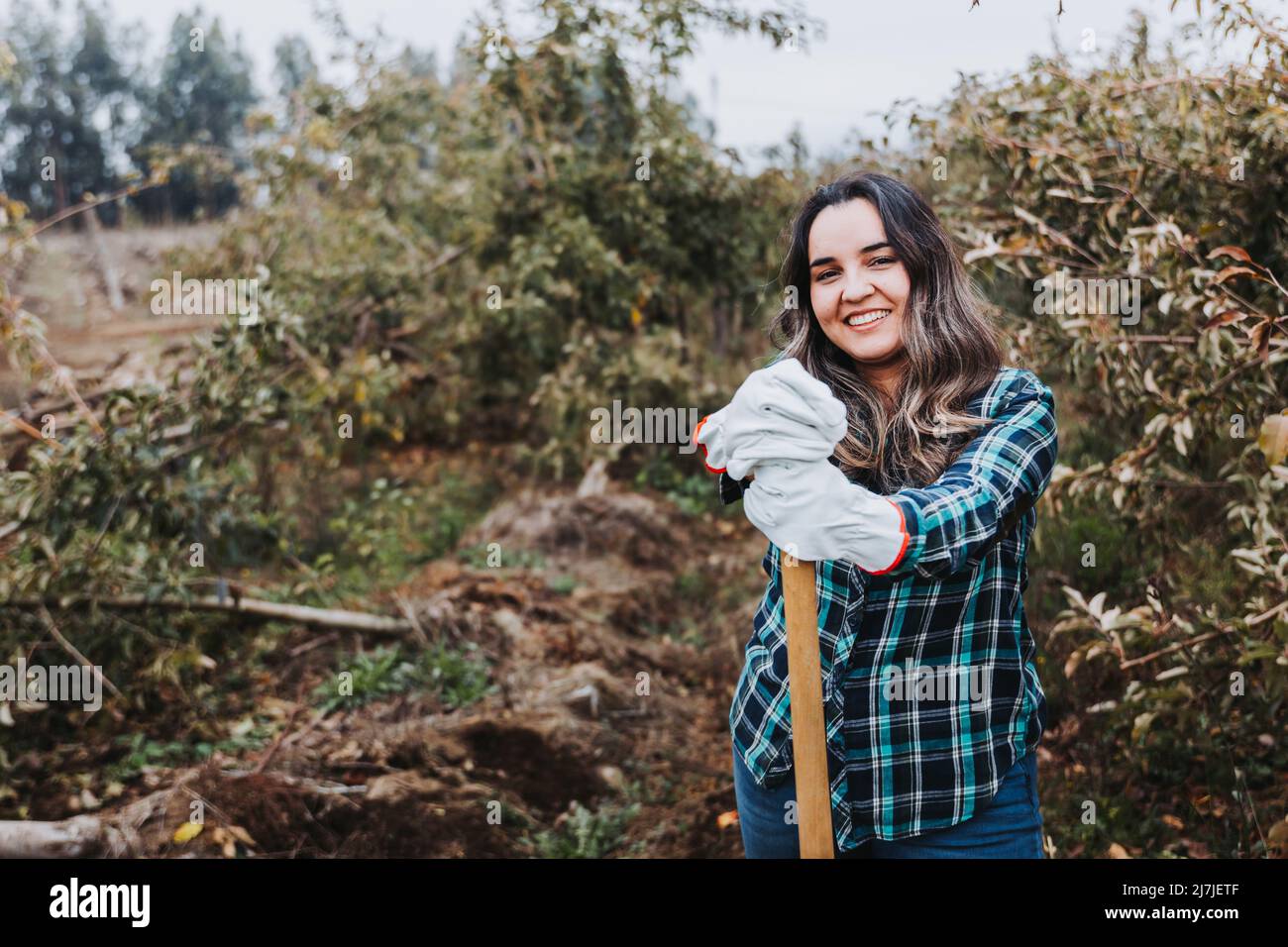 Giovane donna contadina latina sorridente con guanti da giardinaggio e appoggiata su una pala. Foto Stock