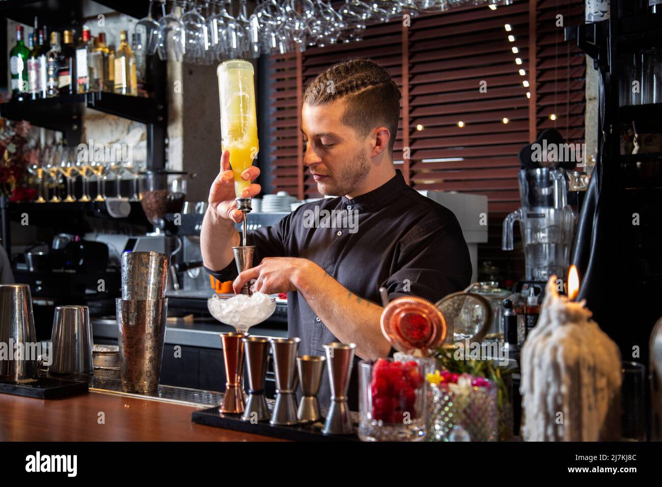 Barman giovane maschio concentrato con acconciatura alla moda in uniforme versare il liquido arancione in jigger mentre si trova al banco e preparare l'alcol c Foto Stock