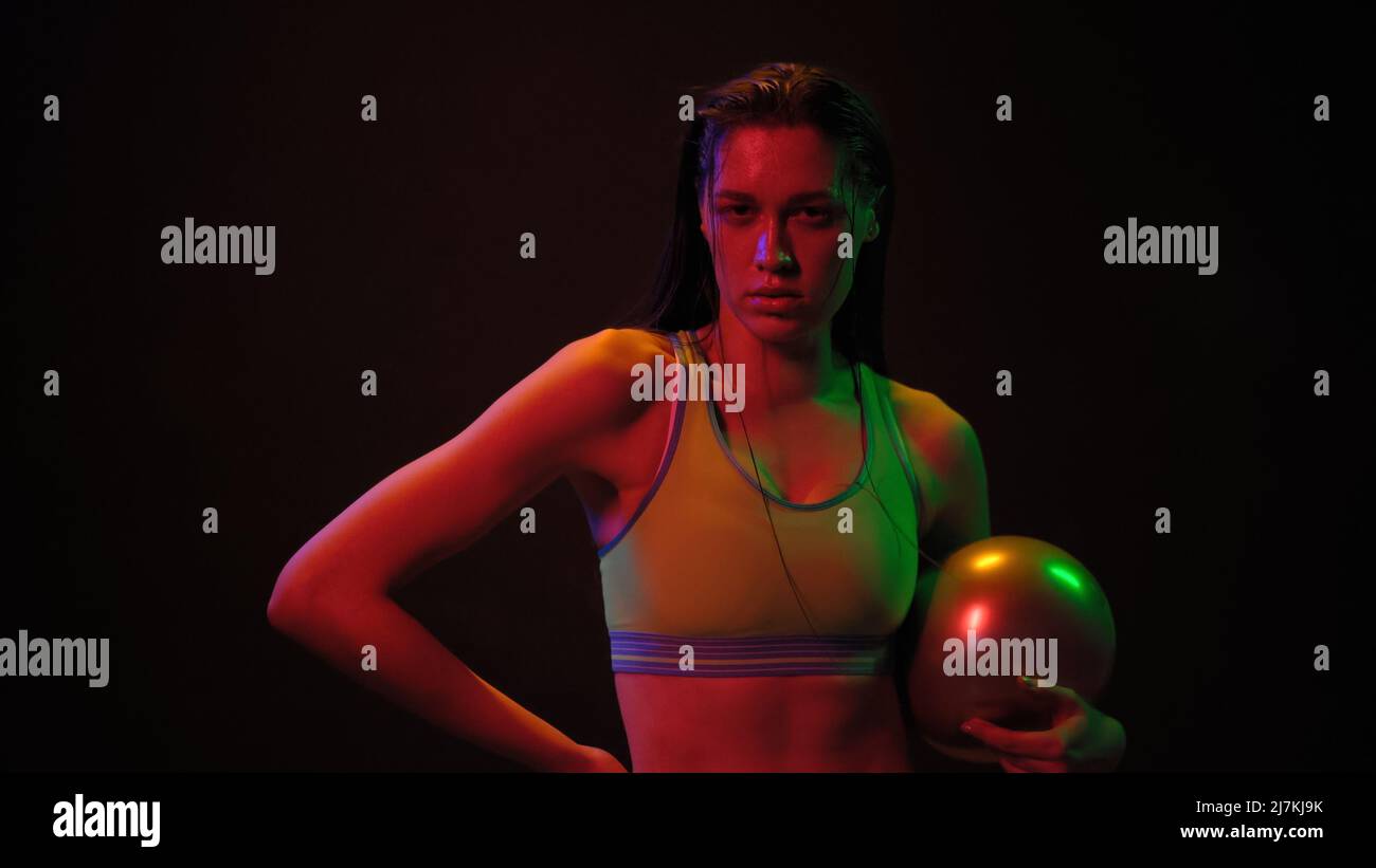 Atleta ragazza con potenza palla esercizio in piedi in luci al neon indossare top sport lavorando su un peso fitness isolato su sfondo nero. Concetto di luce al neon. Foto Stock