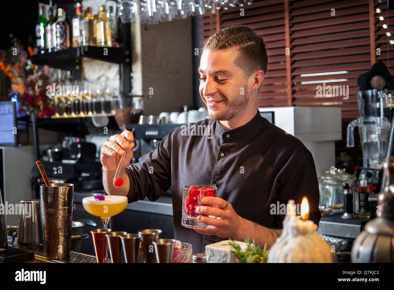 Allegro barman in camicia nera che decora cocktail fresco alcolico aspro  con petali e lamponi mentre si trova al banco con vari strumenti Foto stock  - Alamy