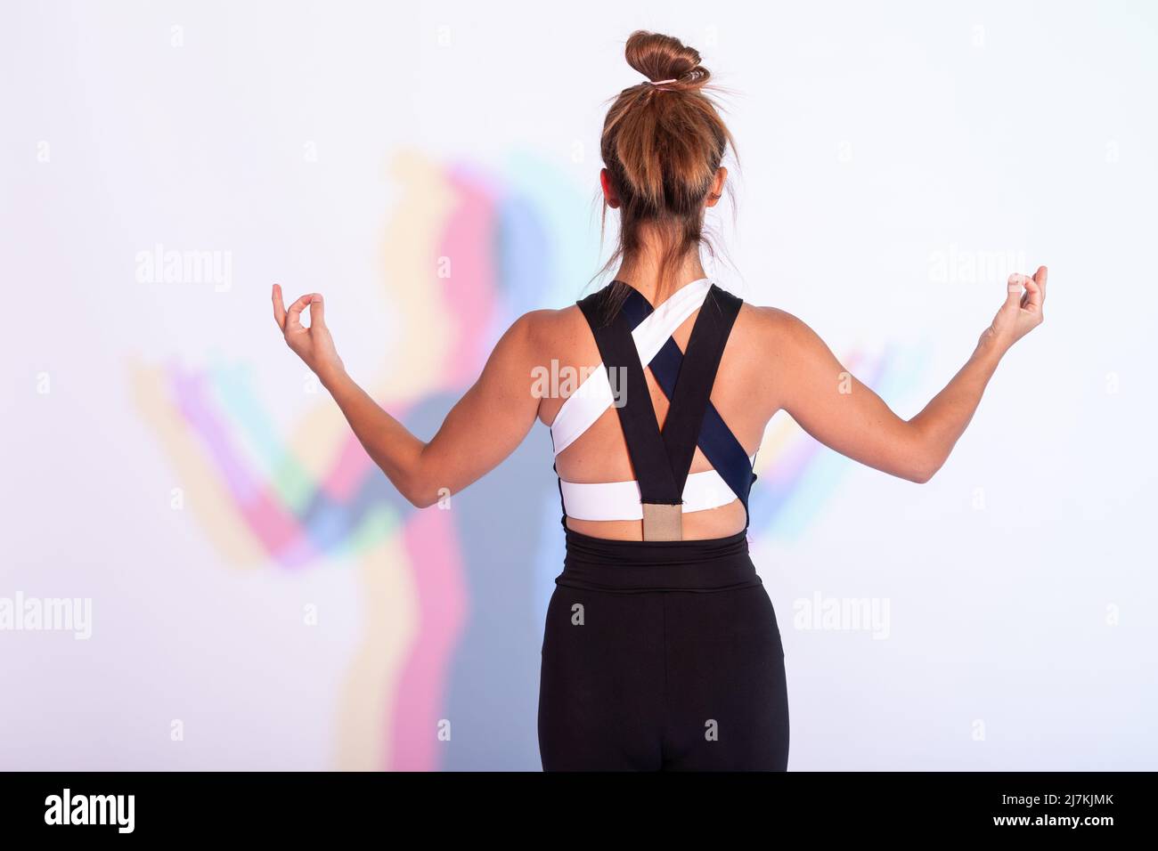 Vista posteriore di una donna irriconoscibile in abbigliamento sportivo meditando con gesti mudra gyan su sfondo bianco con ombre durante l'allenamento yoga Foto Stock
