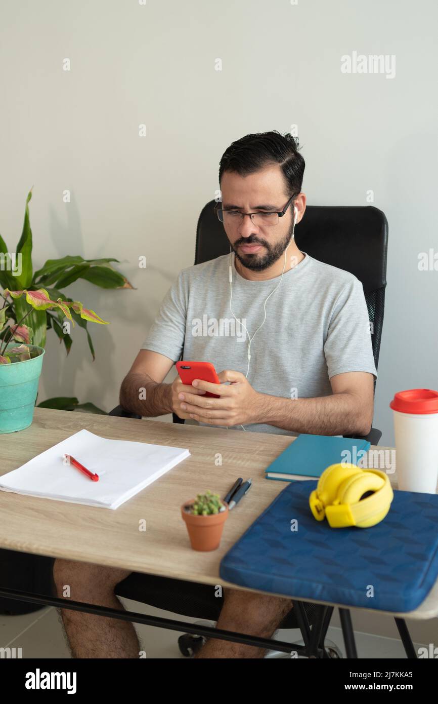 L'uomo si è concentrato nella messaggistica di testo degli auricolari sullo smartphone e nel lavoro di documentazione durante il lavoro a tavola in ufficio Foto Stock
