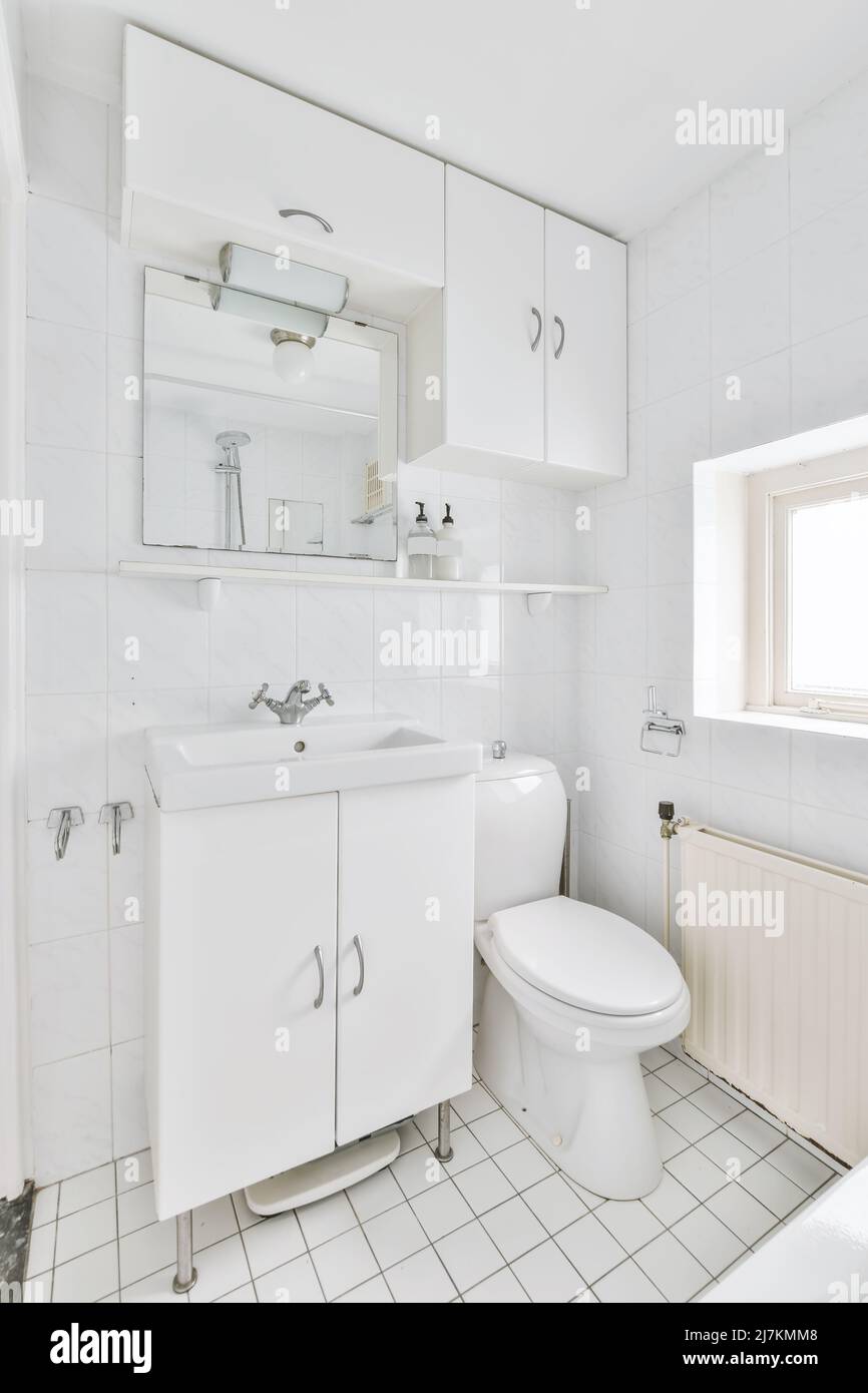 Lavello in ceramica bianca a parete con specchio e prodotti per la cura della pelle posti vicino al gabinetto in bagno luminoso con finestra a casa Foto Stock