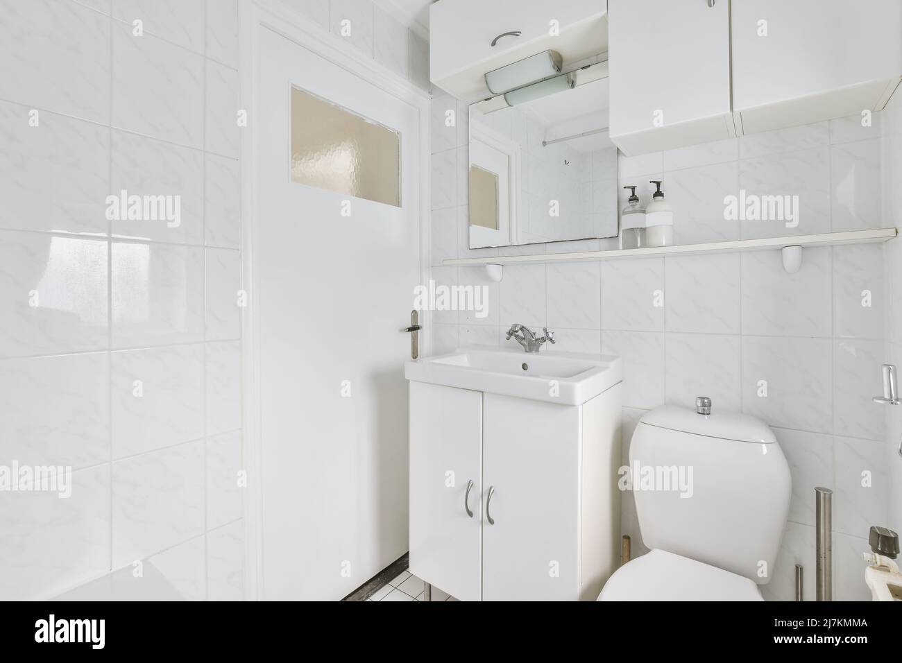 Lavello in ceramica bianca a parete con specchio e prodotti per la cura della pelle posti vicino al gabinetto in bagno luminoso con finestra a casa Foto Stock