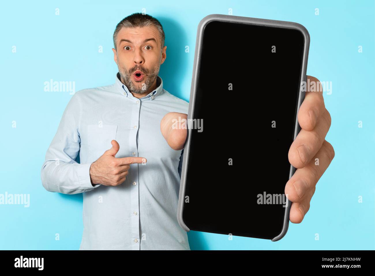 Shocked Male di mezza età che mostra il telefono cellulare schermo, sfondo blu Foto Stock