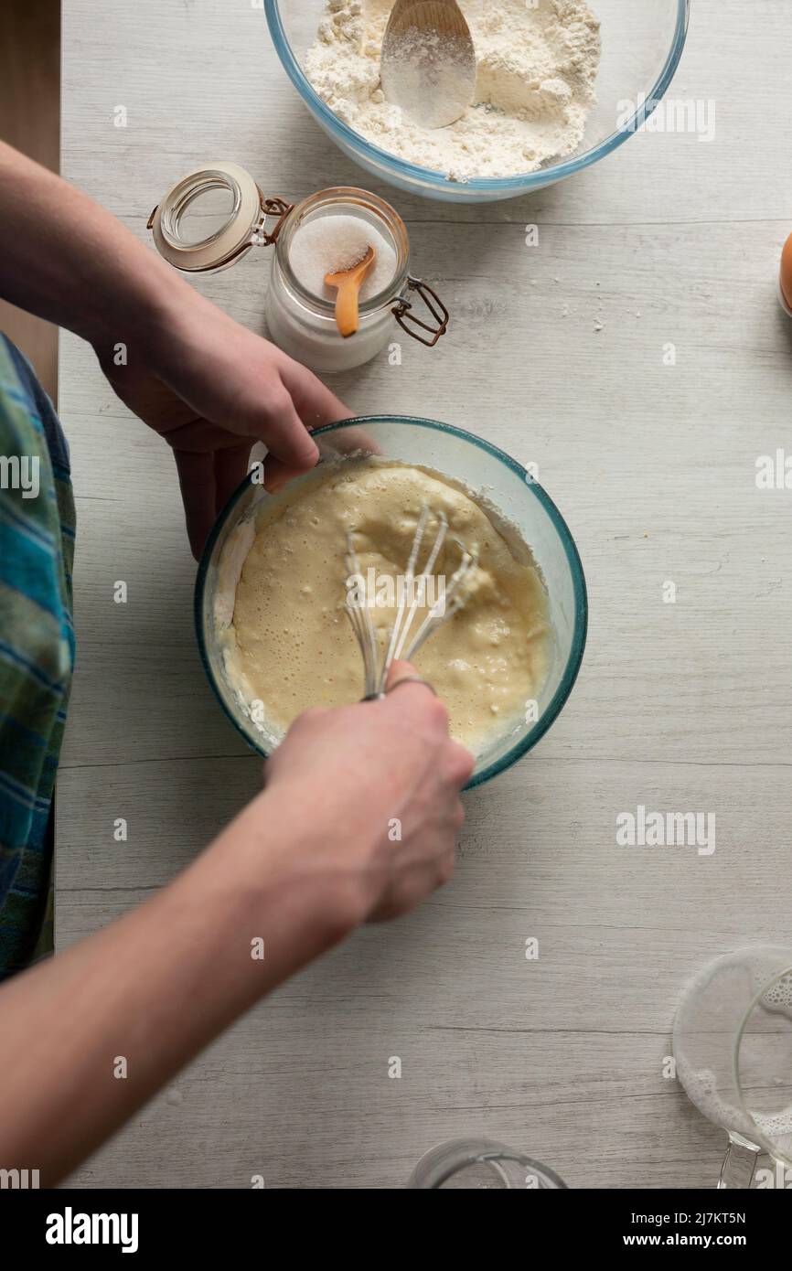 Coltivare persona anonima in pastella di frusta al grembiule per frittelle fatte in casa mentre si cuoce al tavolo con utensili assortiti in cucina leggera Foto Stock