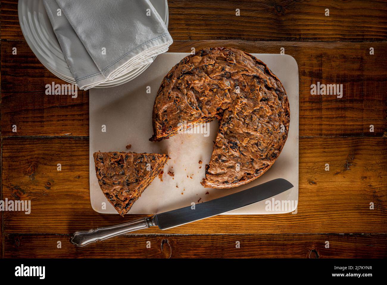 Torta di frutta servita su un pannello di marmo con un segmento tagliato, su un piano di lavoro di legno scuro, con una pila di piatti bianchi e tovaglioli sullo sfondo. Foto Stock