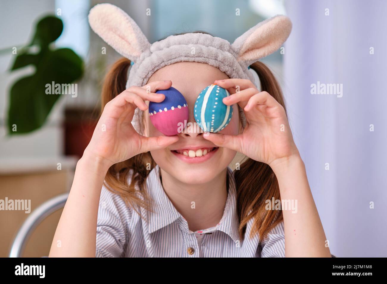 Una ragazza giocosa positiva in orecchie conigliere tiene le uova colorate nelle sue mani nella cucina domestica. Buona Pasqua. Sfondo di Pasqua Foto Stock