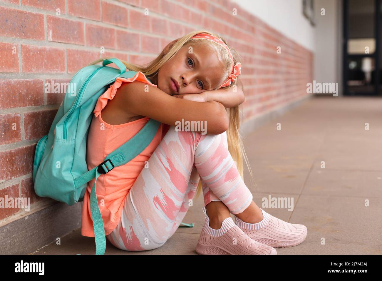 Ritratto di scolaretta elementare caucasica con zaino seduto sul pavimento nel corridoio della scuola Foto Stock