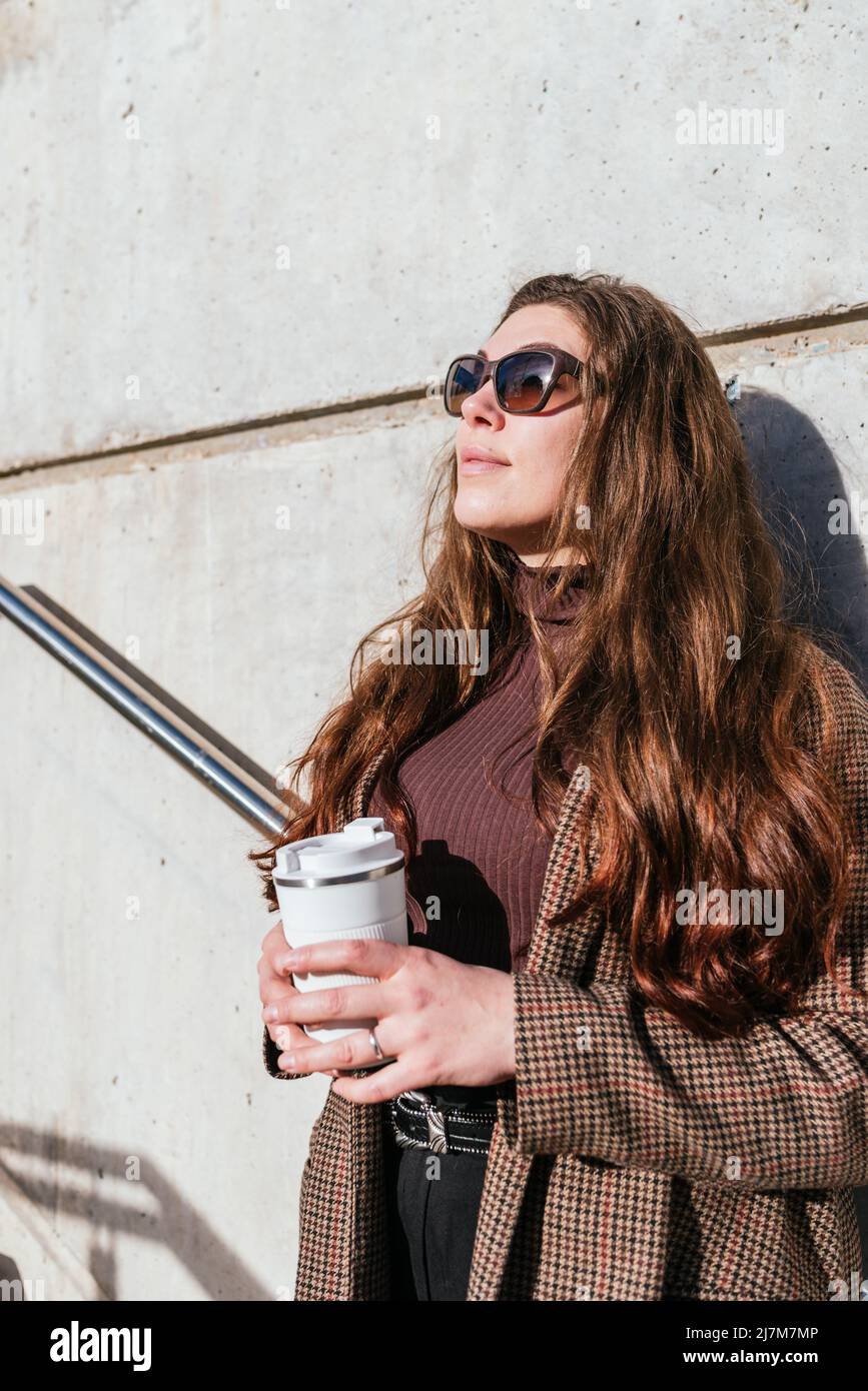 Donna elegante in outerwear con occhiali da sole appoggiati a parete e guardando via mentre si gusta il caffè per andare e sognare sulla strada della città Foto Stock